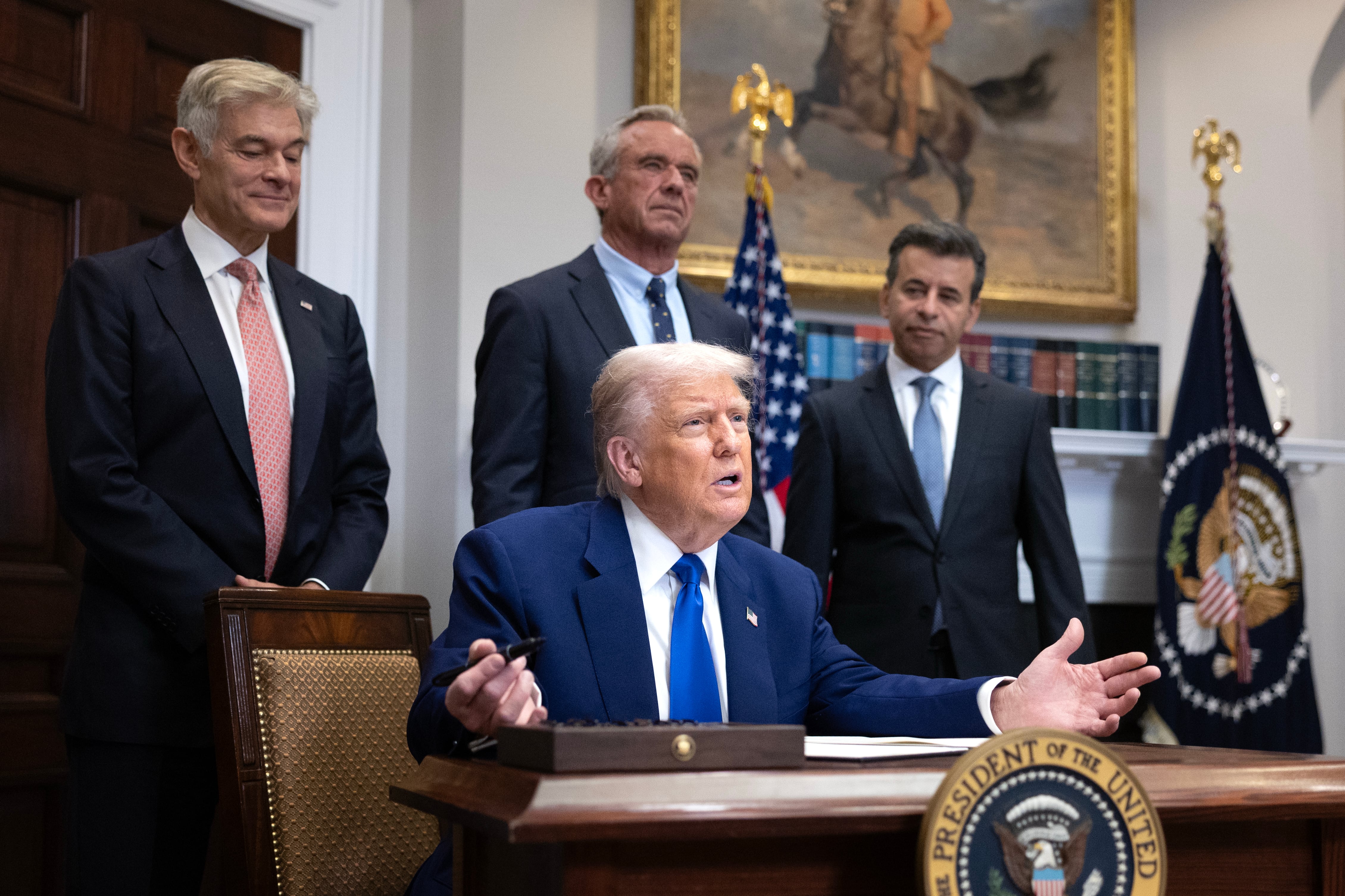 President Donald Trump is seated behind a desk, with Health and Human Services Secretary Robert F. Kennedy Jr., National Institutes of Health Director Jay Bhattacharya, and Dr. Mehmet Oz, Administrator for the Centers for Medicare & Medicaid Services, standing behind him.