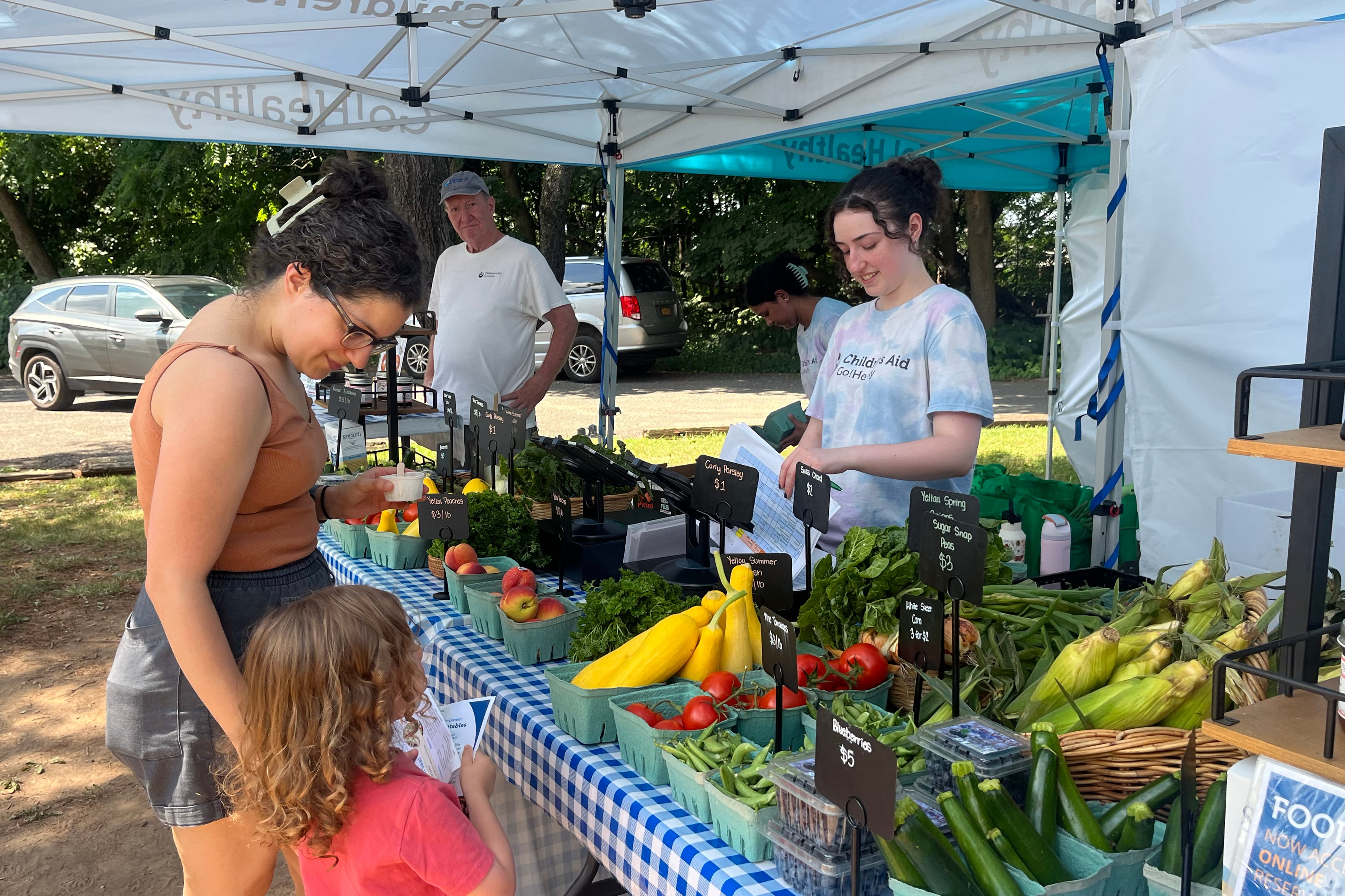 A photograph of a mother and her young son standing on one side of a table full of fresh produce while two people stand on the other side all under a large tent and a car in the background.