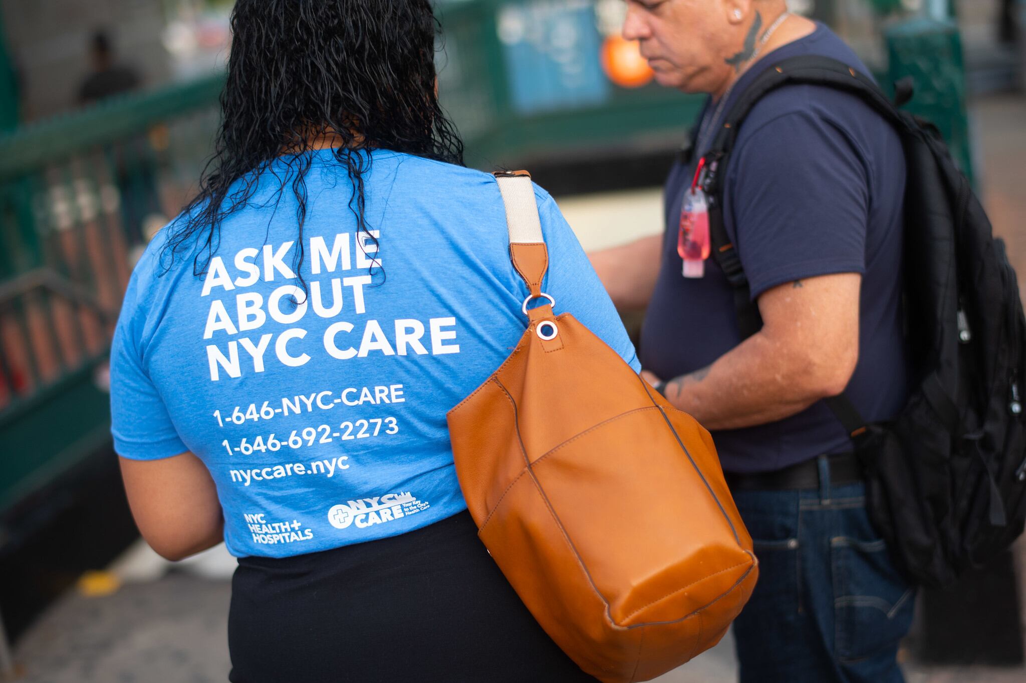 Two people confer on the street, one wearing a T-shirt that says "Ask me about NYC Care."