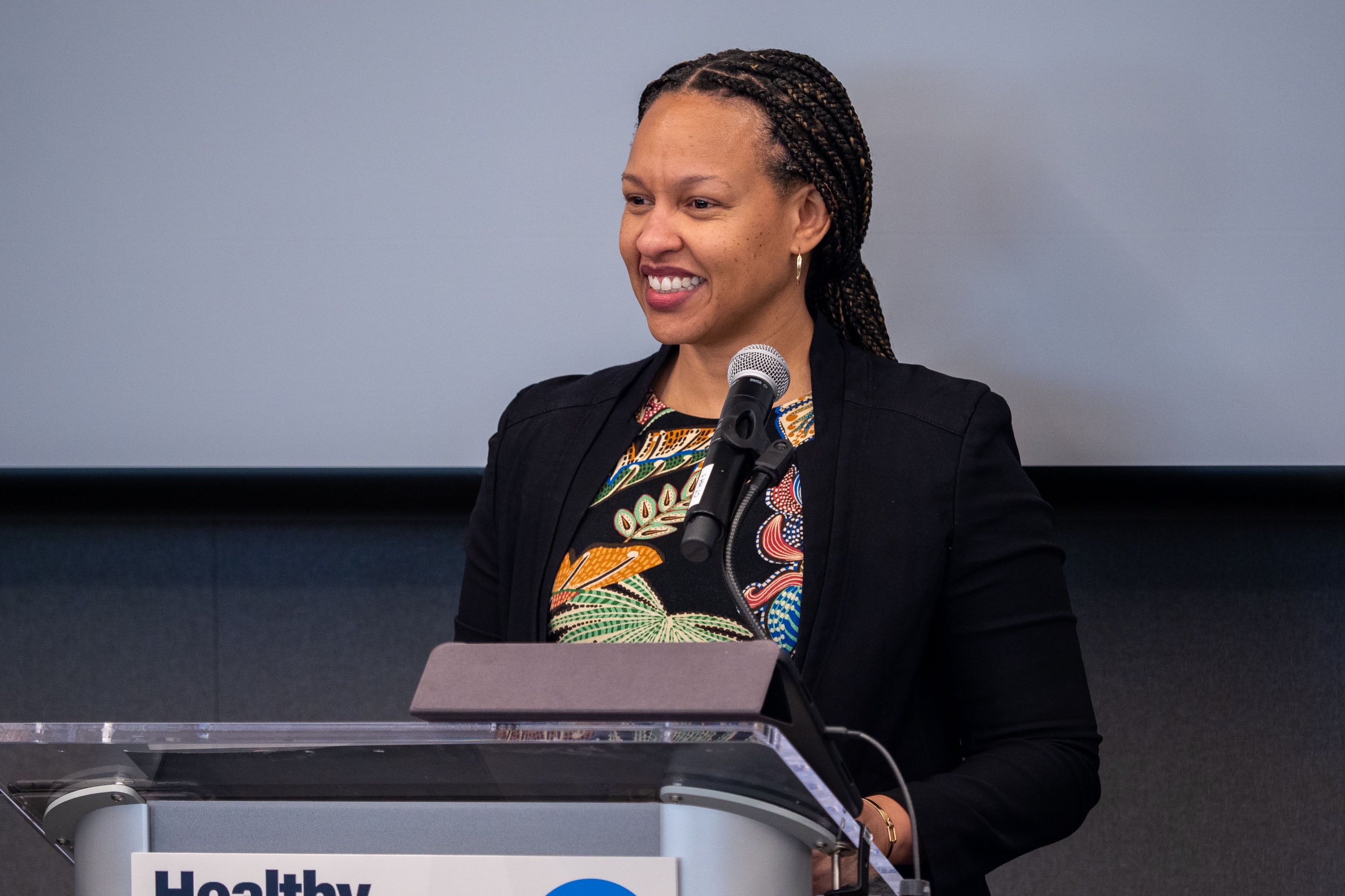 A Black woman with long dark hair and wearing a suit jacket speaks from a podium in a conference room.