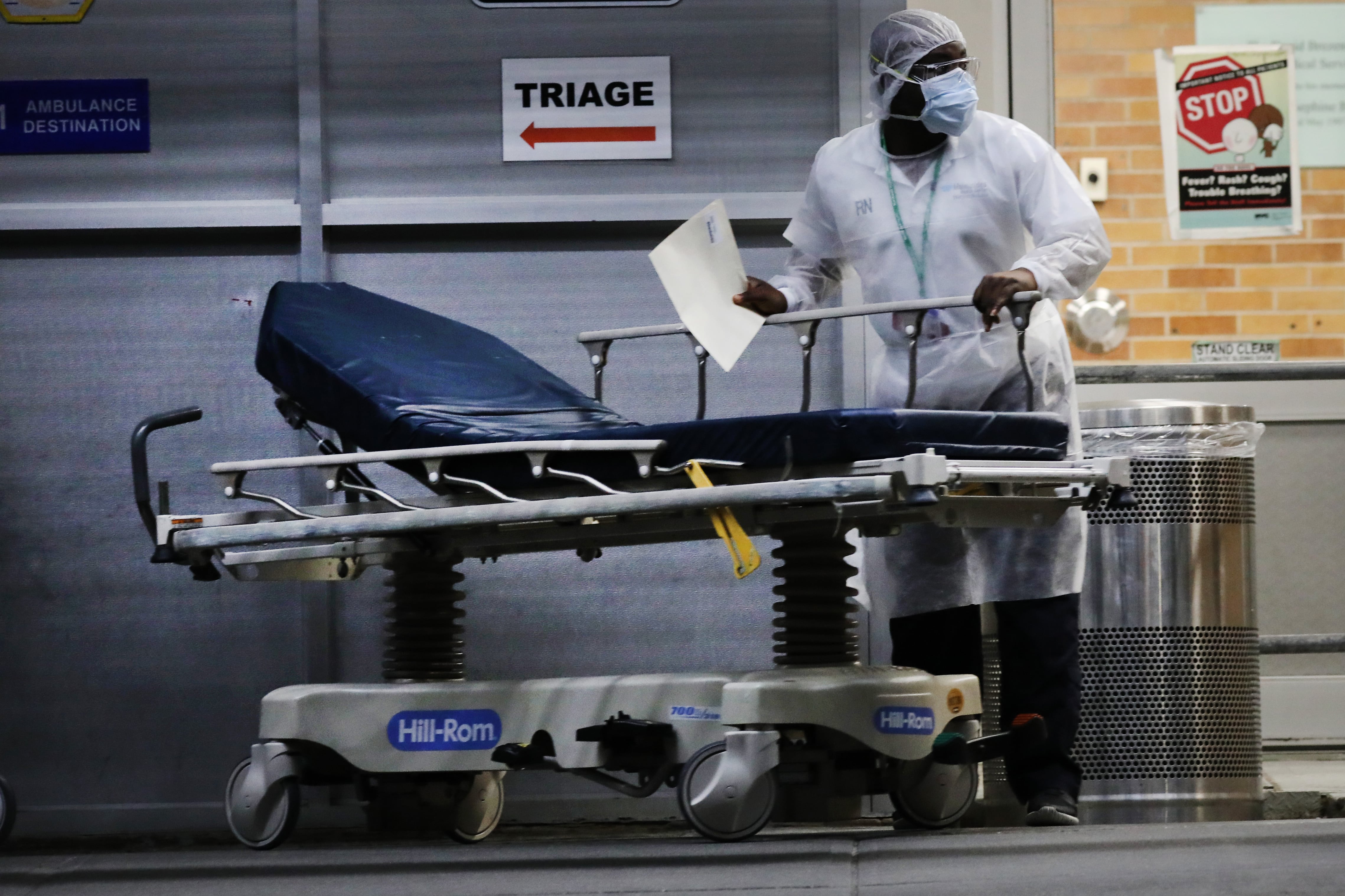 A medical worker wearing a face mask and protective gear stands with a gurney outside a hospital.