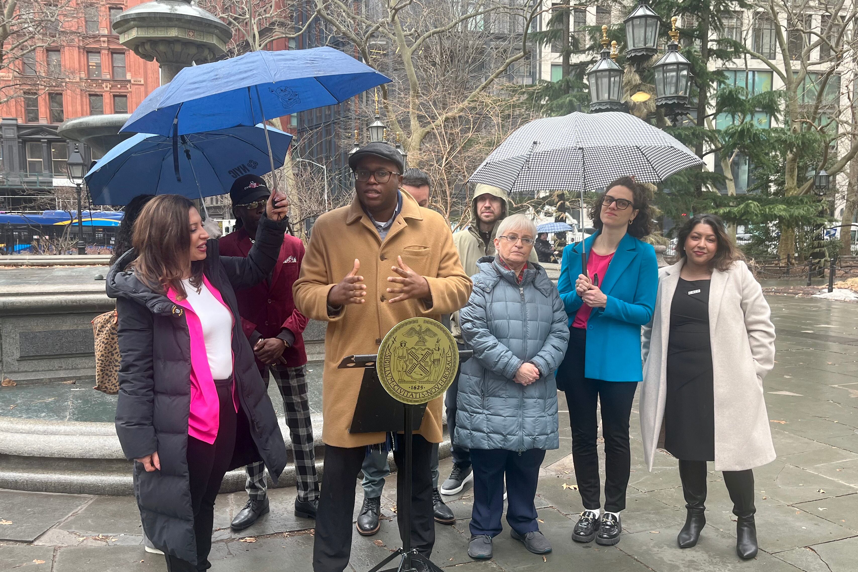 A group of people in coats and holding umbrellas gather outside and speak in front of a small podium in front of a large group of buildings.