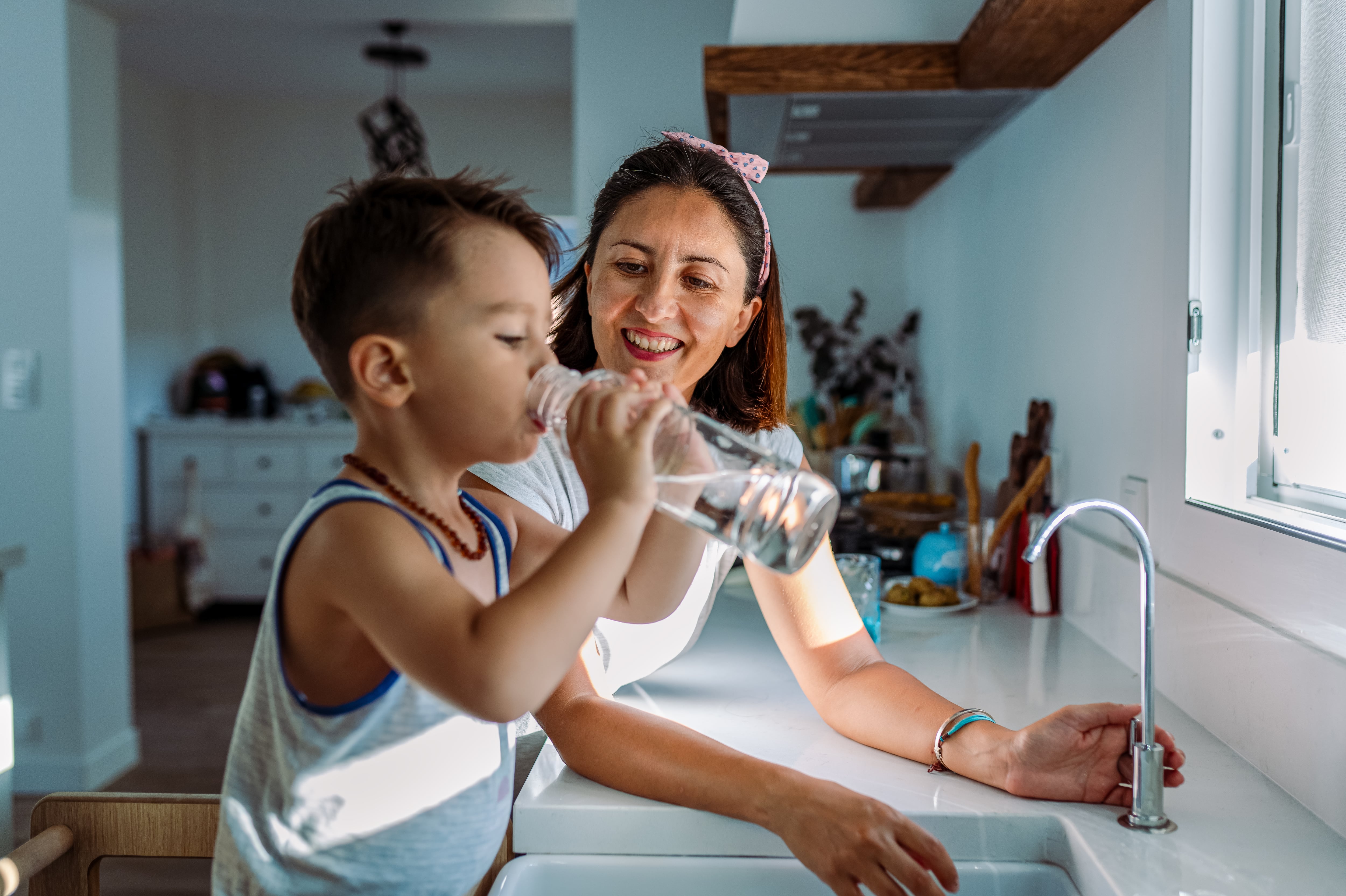 A mom and child stand at sink as child drinks tap water from a bottle.