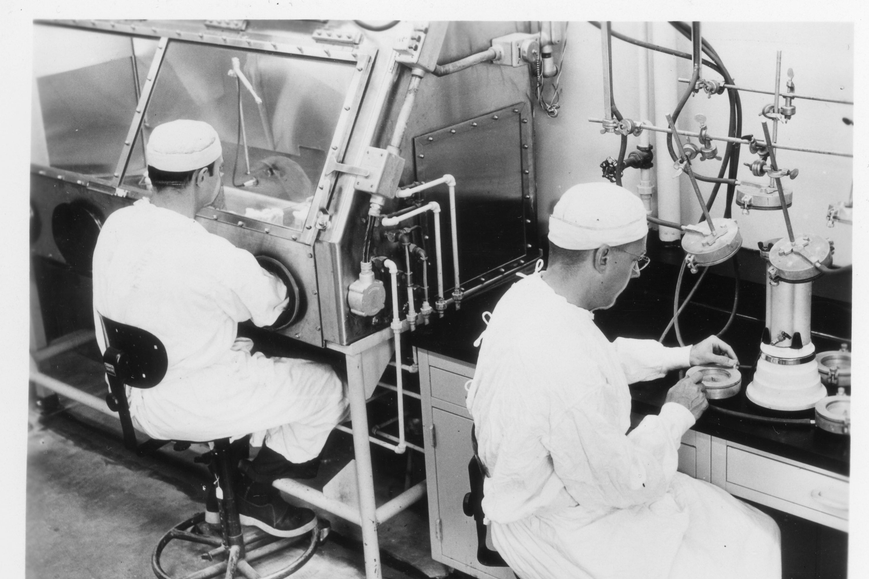 Two men in white caps and laboratory gowns. One man is working on a black desktop with several devices. The other man has his arms inserted in a metal and glass biological safety cabinet.