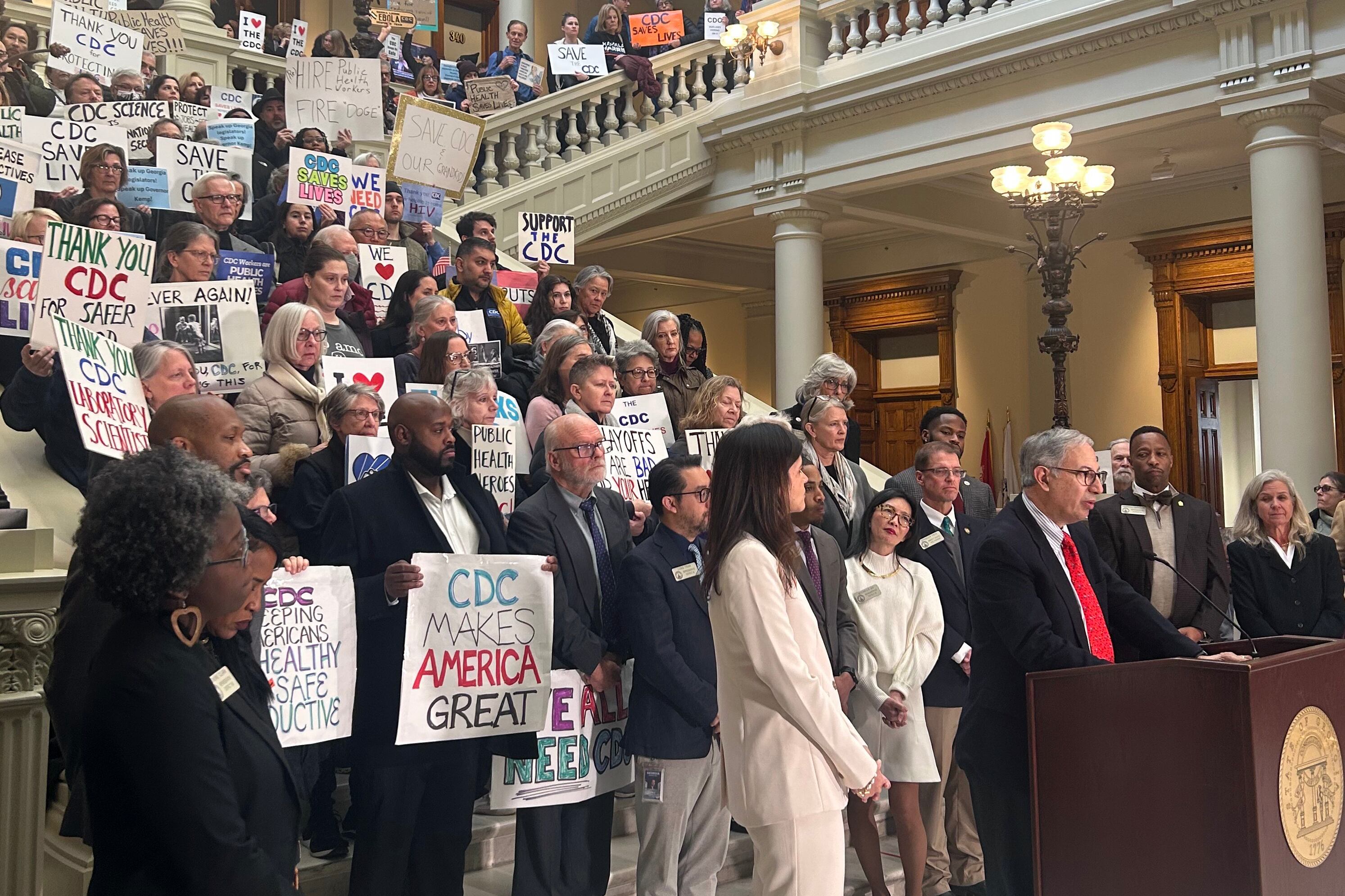 A group of people in suits and business attire, some holding signs in a large crowd on the steps inside of a Capitol building.