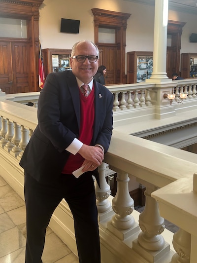 A white man in a suit smiles for a portrait overlooking an open area in a capitol building.