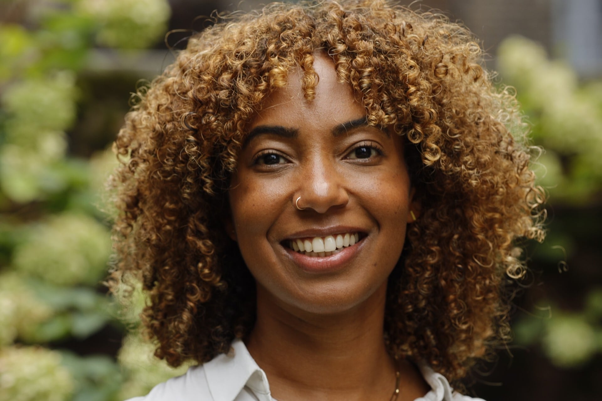 A Black woman with light brown curly hair poses for a portrait in a white blouse outside with some greenery in the background.