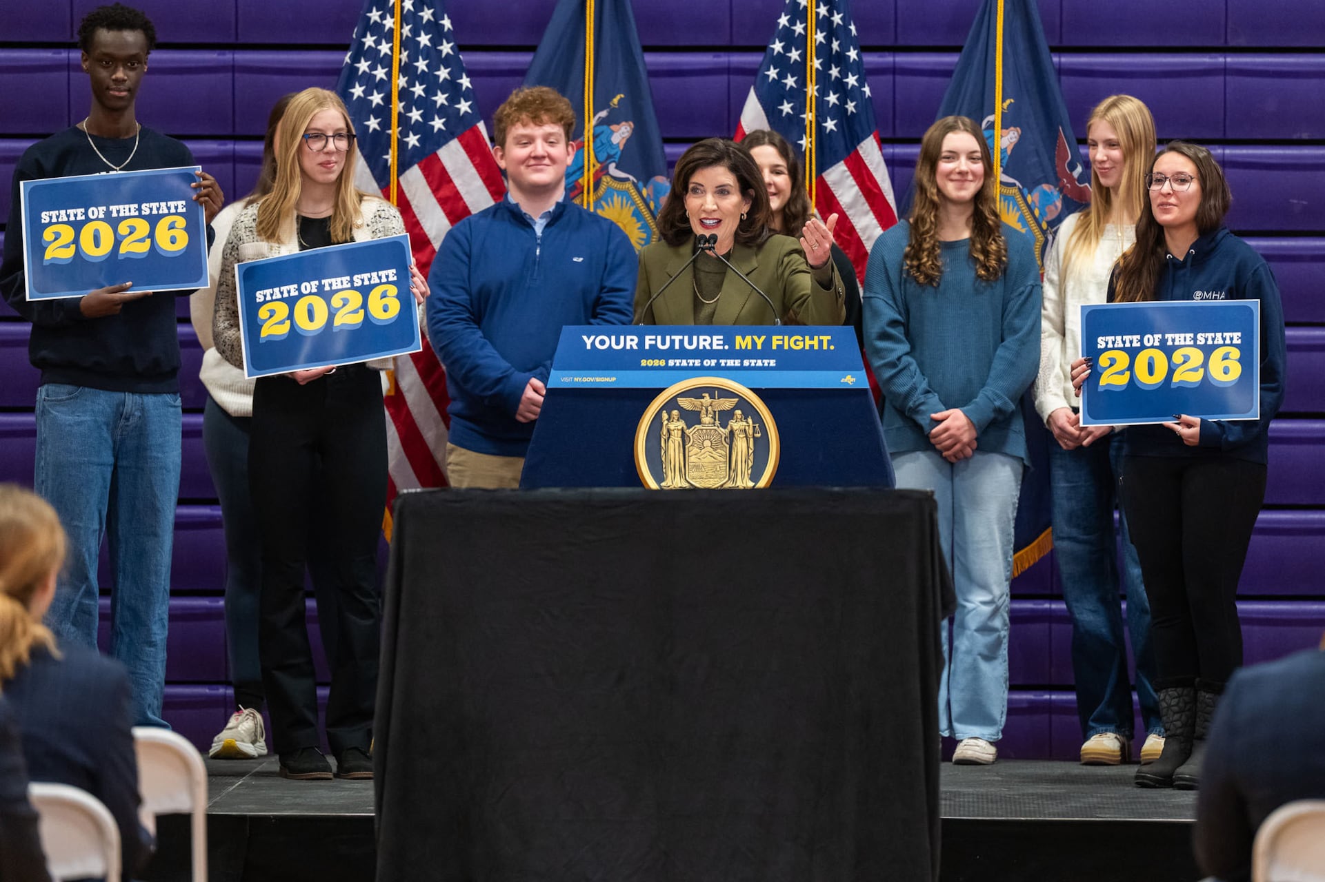 A photograph of a woman in a suit standing behind a podium with a group of people standing behind her.
