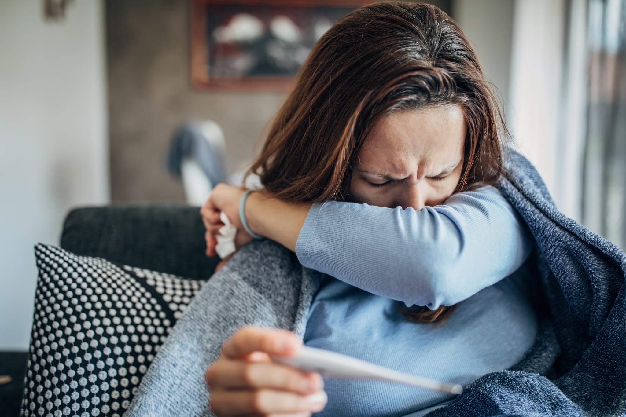 A woman on a couch in a blue long-sleeve shirt holds a tissue and a thermometer as she coughs into her arm.