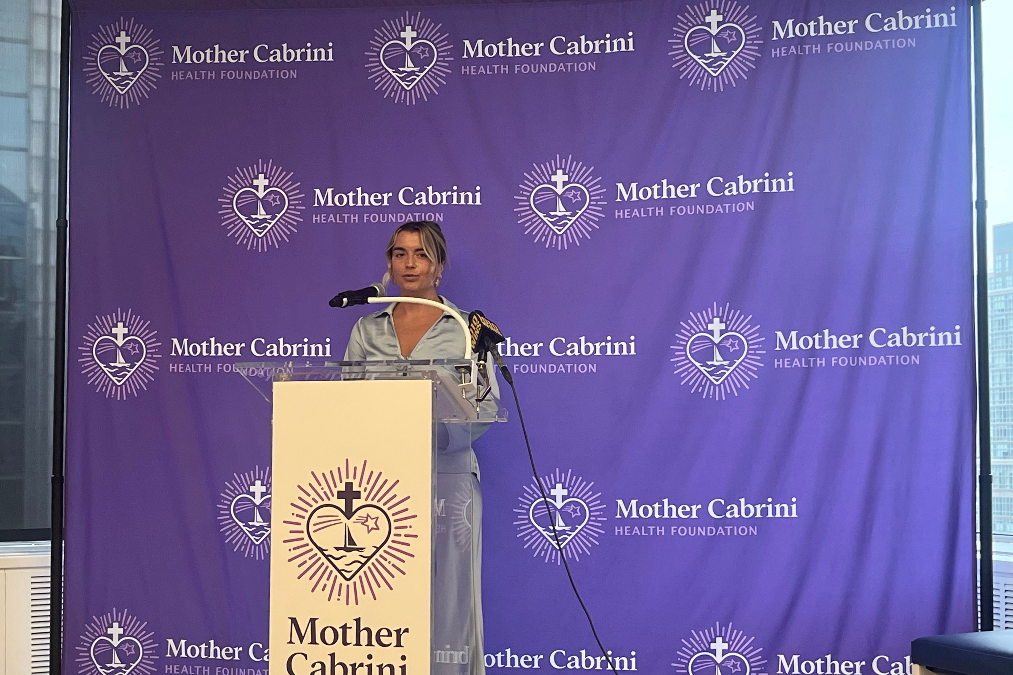 A woman stands behind a podium and in front of a purple backdrop for a press conference.