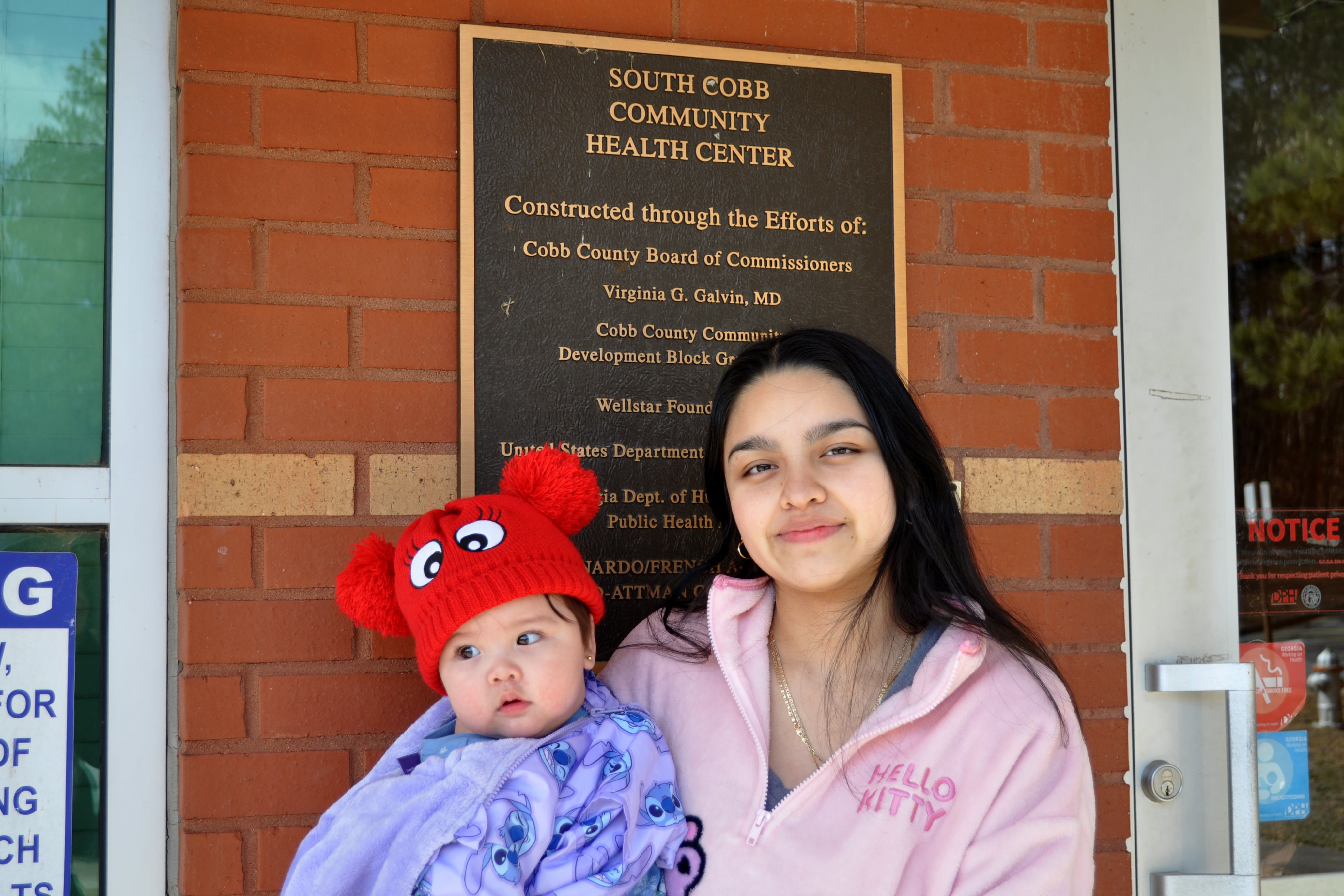 A young woman wearing a pink jacket holds a young baby in front of a red brick building.