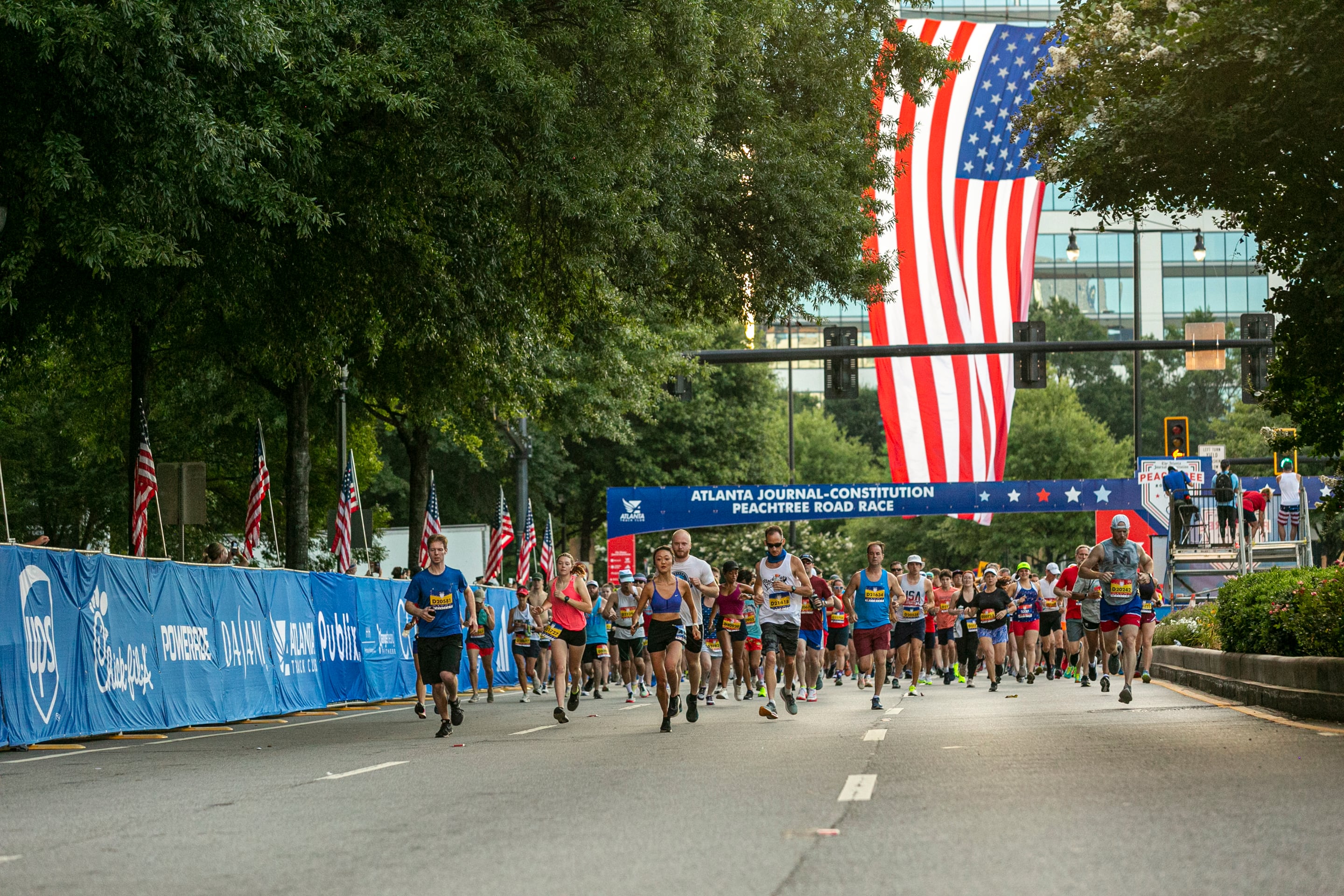 A group of runners crosses the start line at a road race backed by a large American flag.