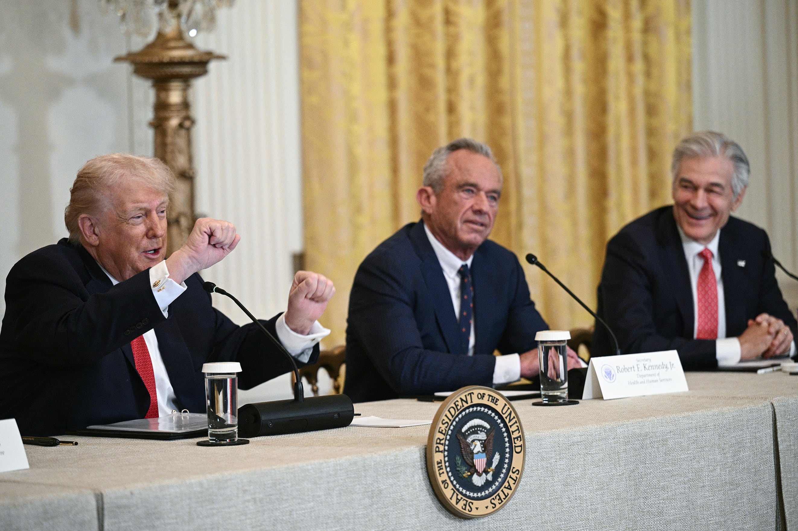 Three white men in suits sit at a table with microphones in a conference room in the White House.