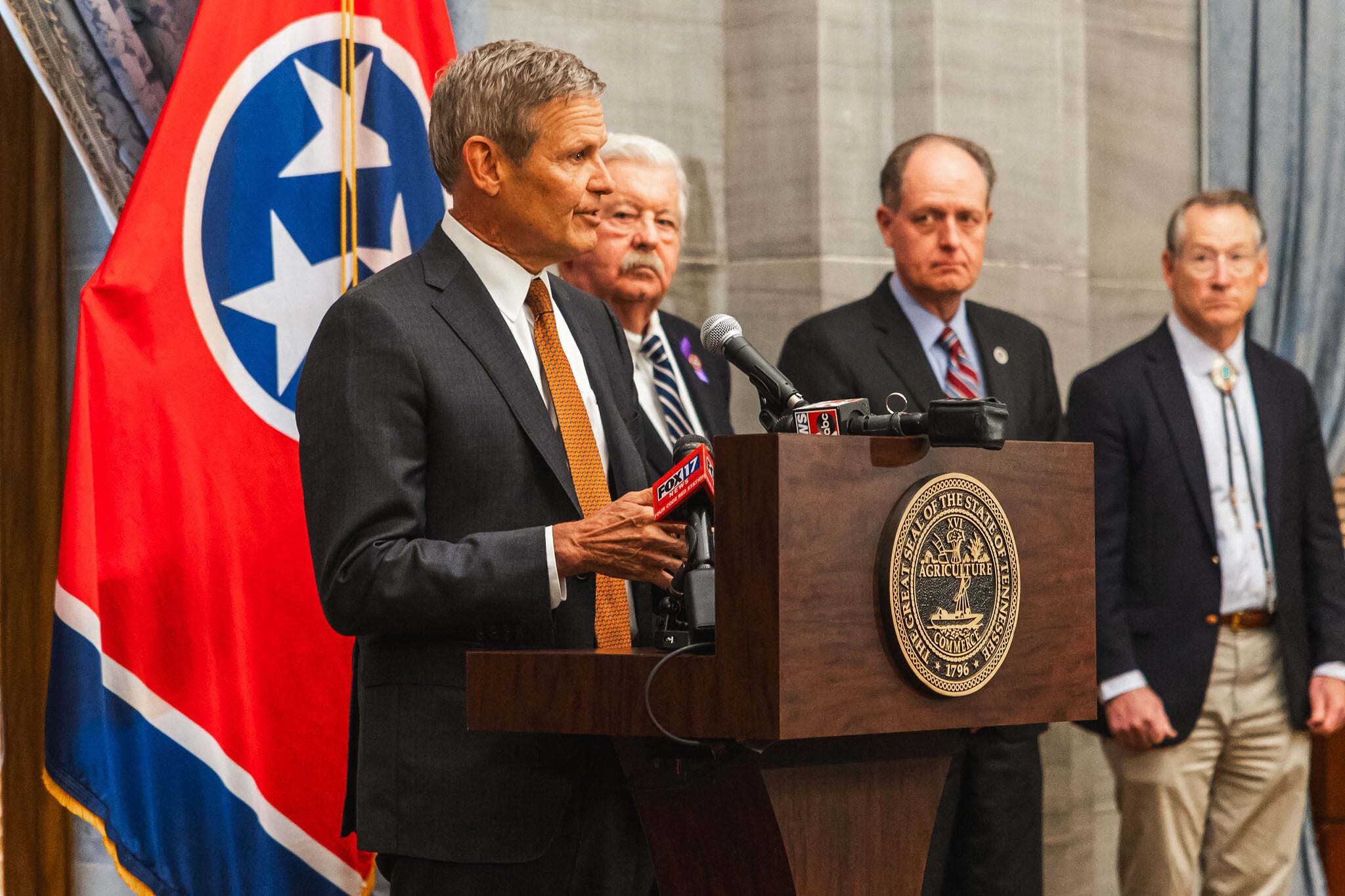 A man in a dark suit stands behind a wooden podium with a Tennessee state flag and three men standing in the background.