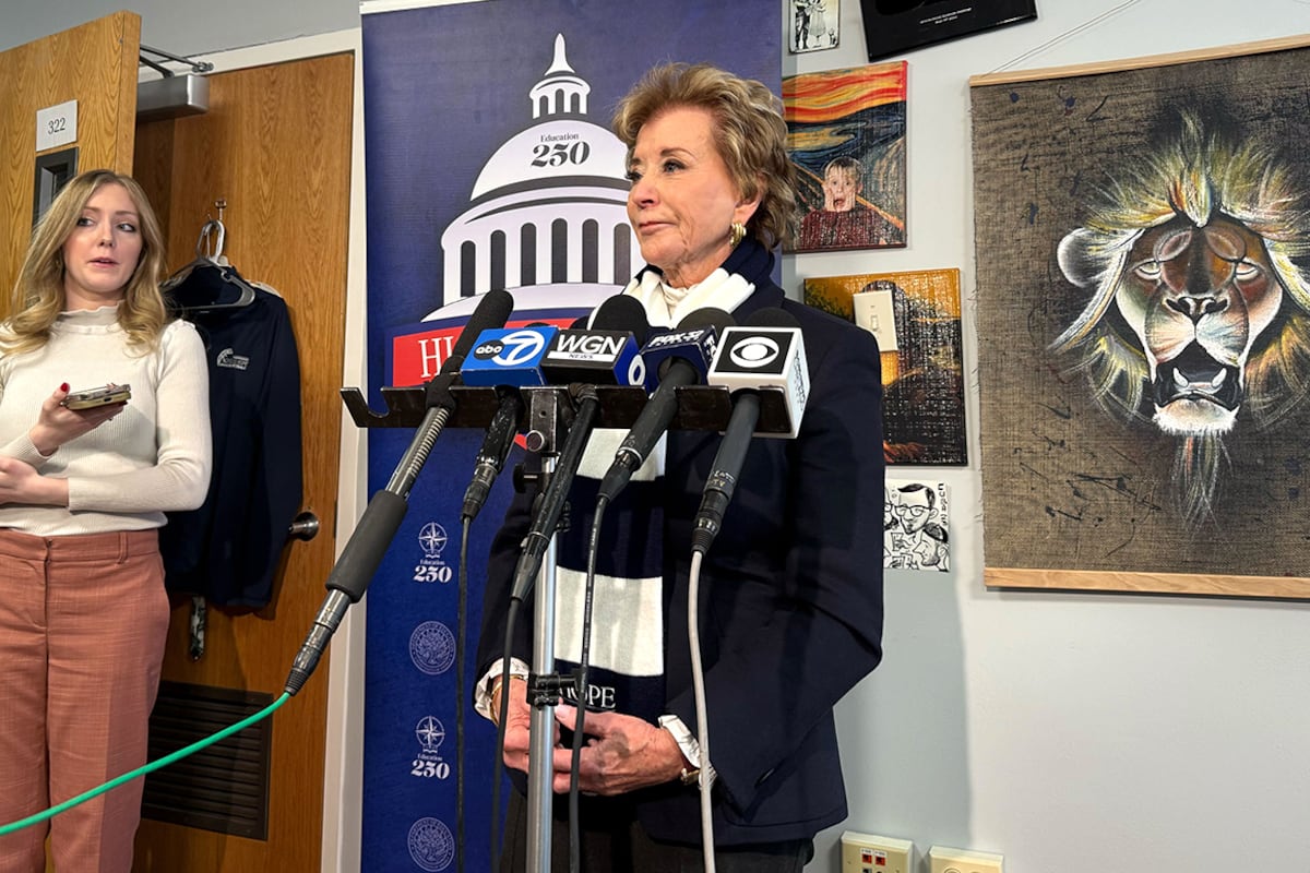 A photograph of a white woman in a suit speaking from behind a podium in a classroom with a wall full of posters and signs in the background.