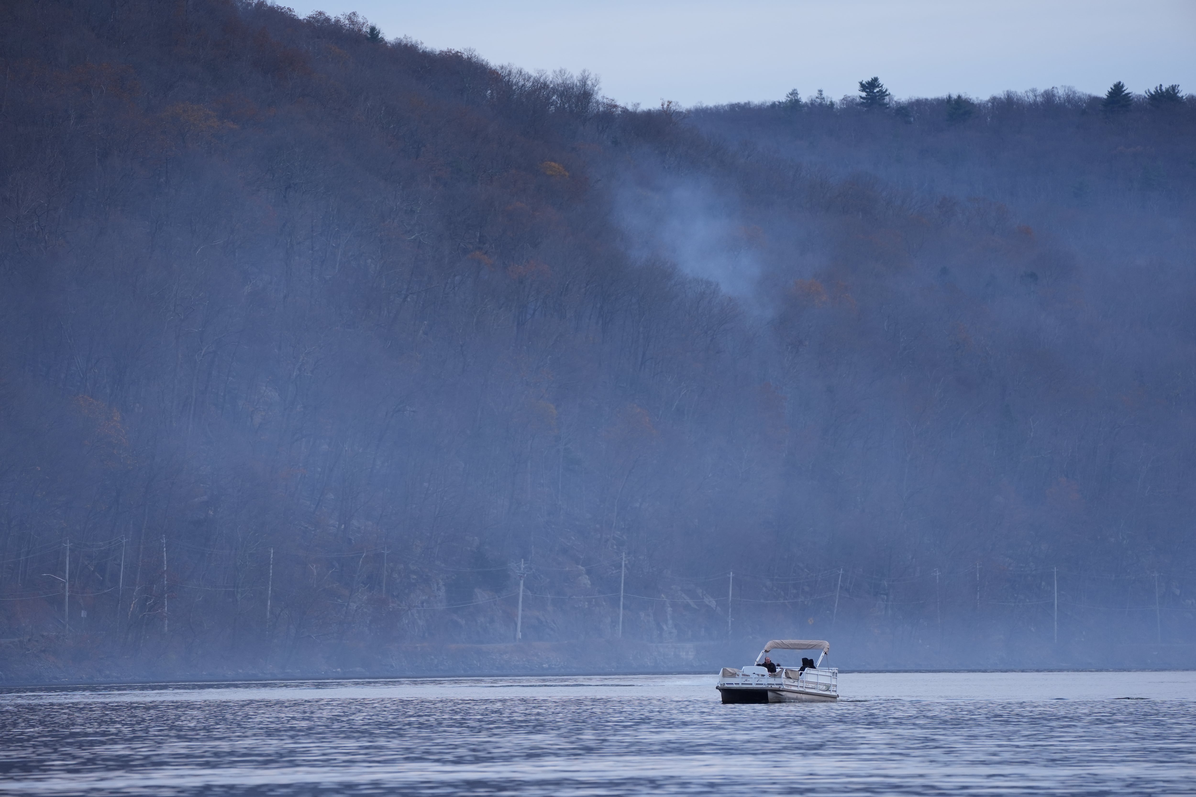 A boat sails on water offshore where a wildfire is burning, sending heavy smoke into the air.