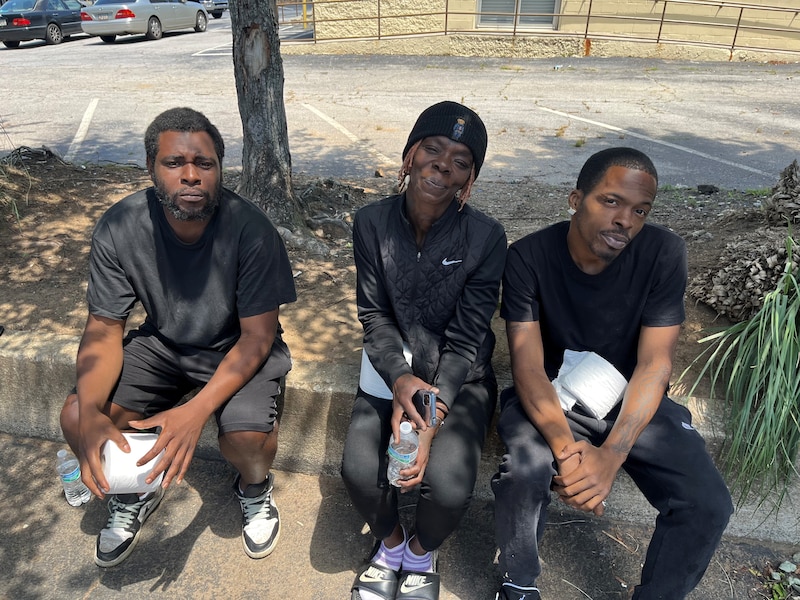 Three people sit on a sidewalk outside an extended-stay hotel.