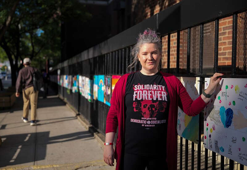 A woman stands in a black t-shirt and burgundy sweater next to a fence. (This is a cropped version of the original.)