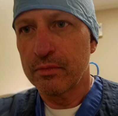 A man wearing a surgical cab and a blue "scrub" top looks into the camera.