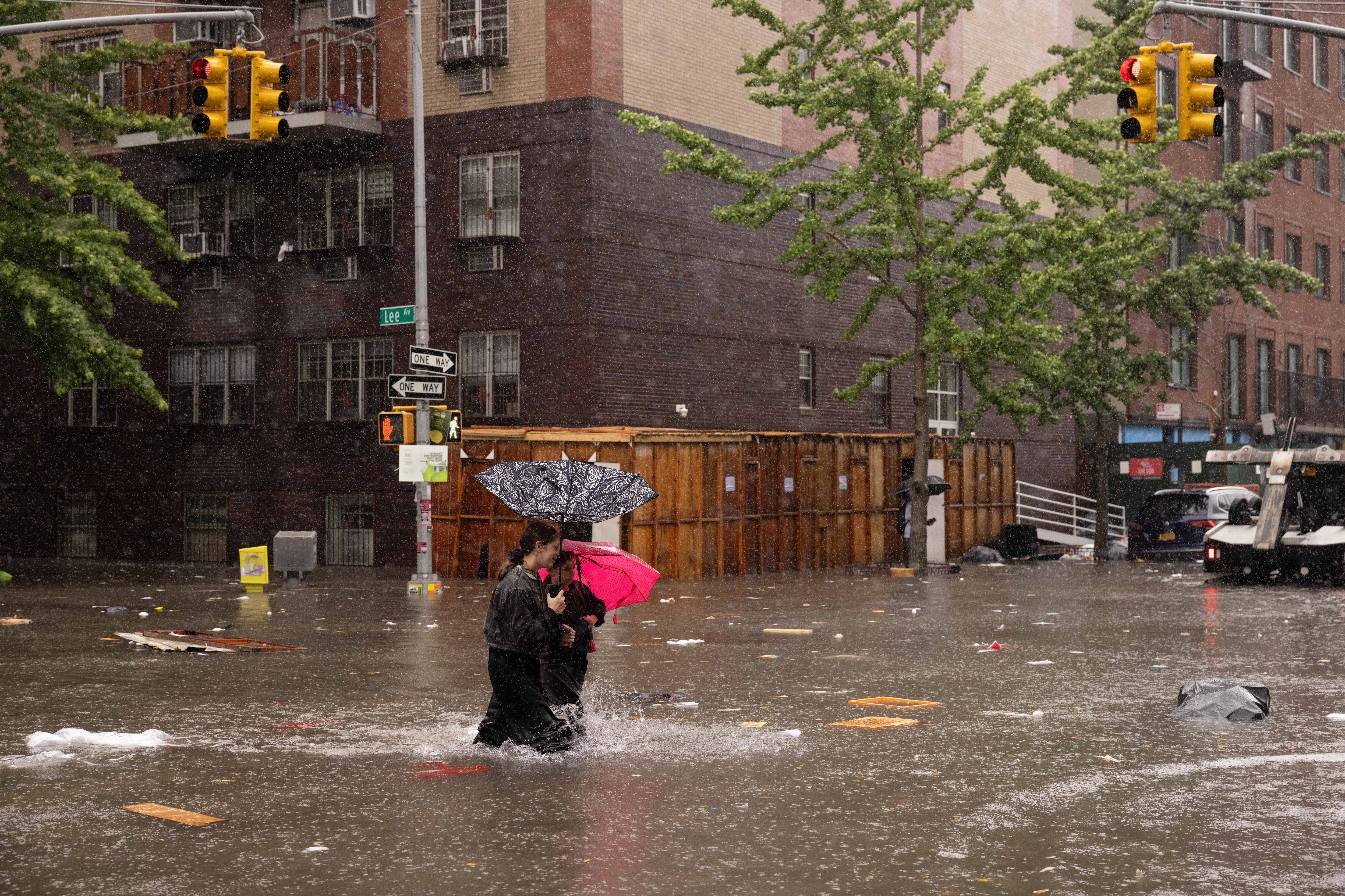 Two people holding umbrellas walk through thigh-high water on a flooded New York City street.