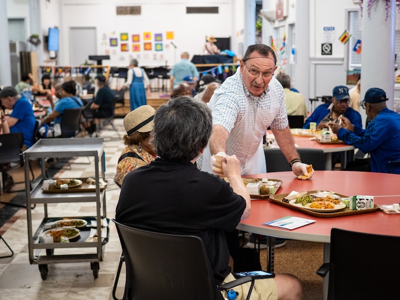 A man in white clothes and a hairnet hands out food at a table.
