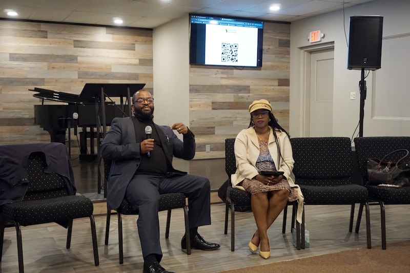 Two people talk while sitting in a row of chairs in a church building.