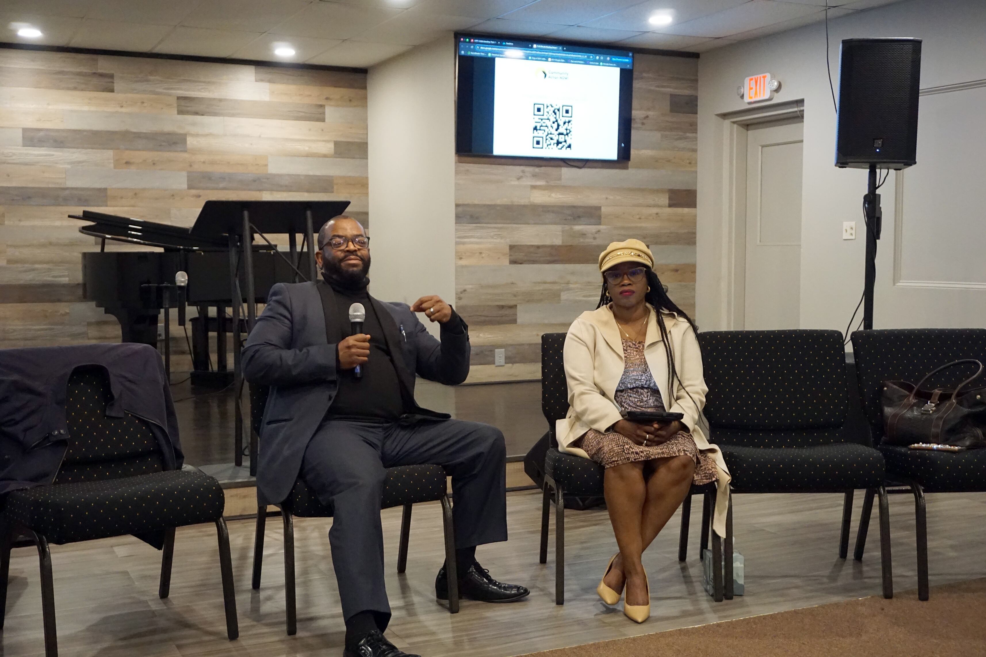 Two people talk while sitting in a row of chairs in a church building.