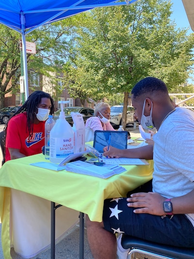 Two Black men sit at a table outside wearing masks.