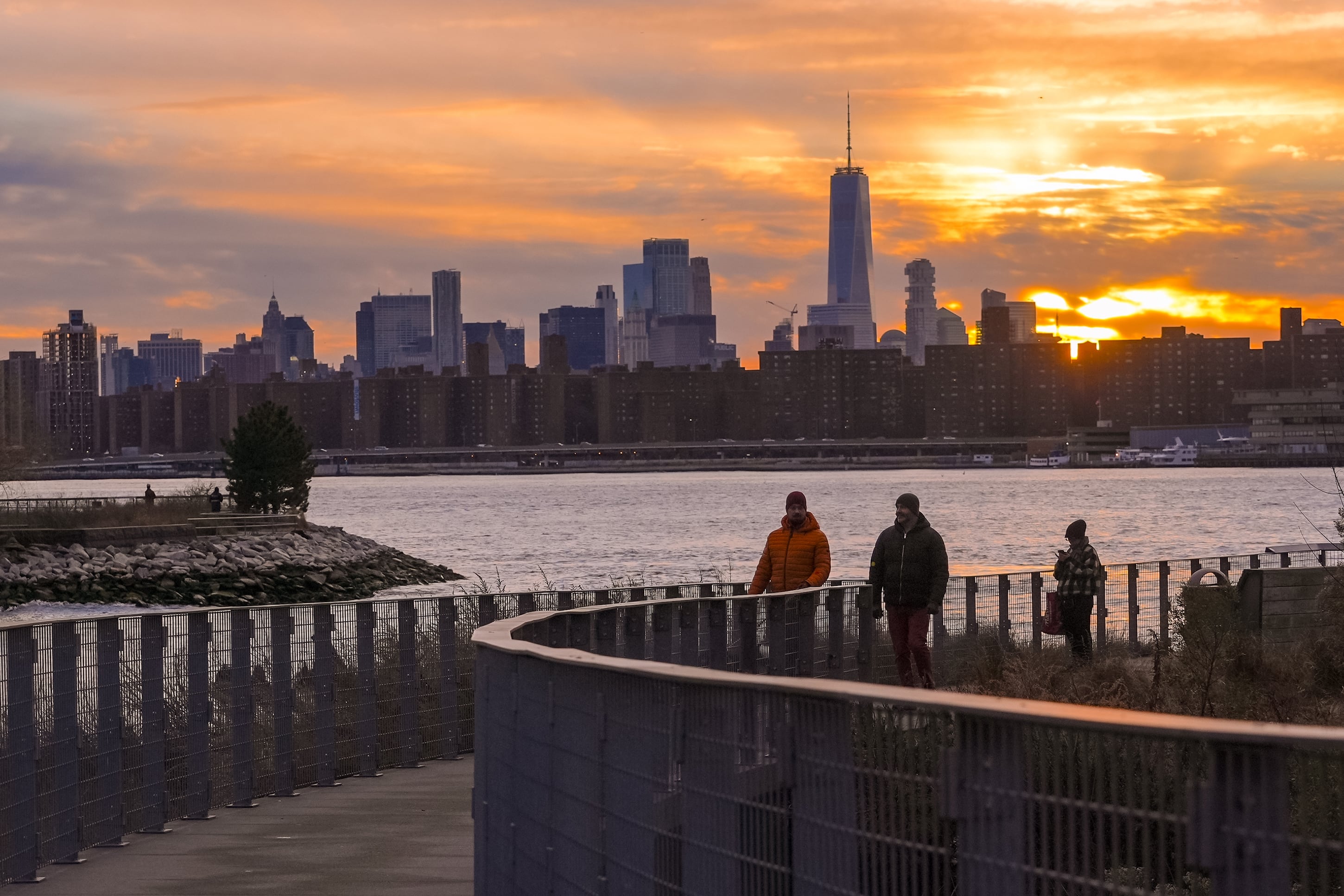Three people in coats walk along a river with a large cityscape in the background while the sunsets.