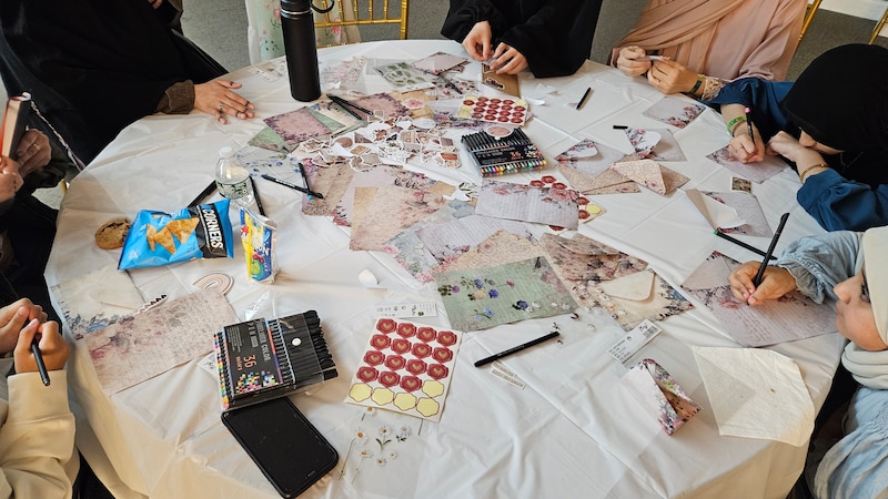 Women and girls write and decorate letters at a round table.