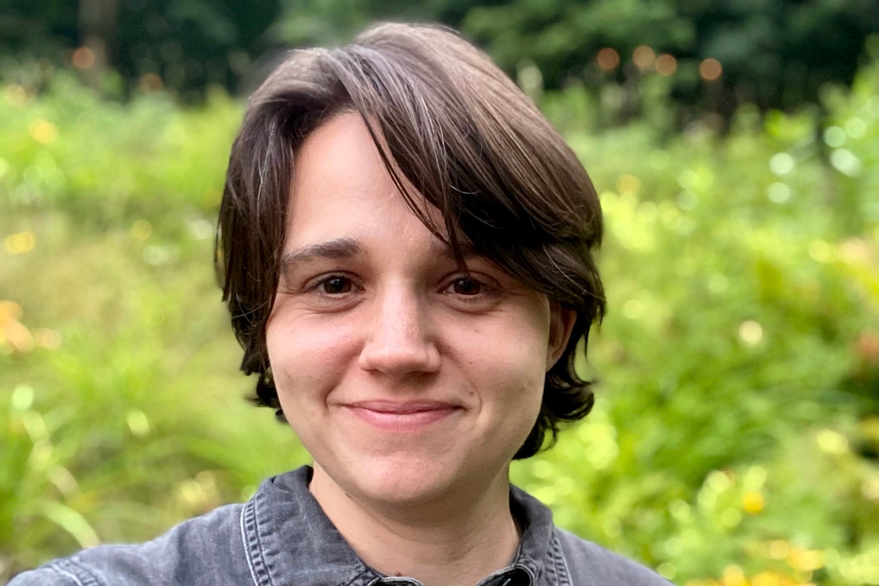 A person with short dark hair smiles at the camera with greenery in the background.