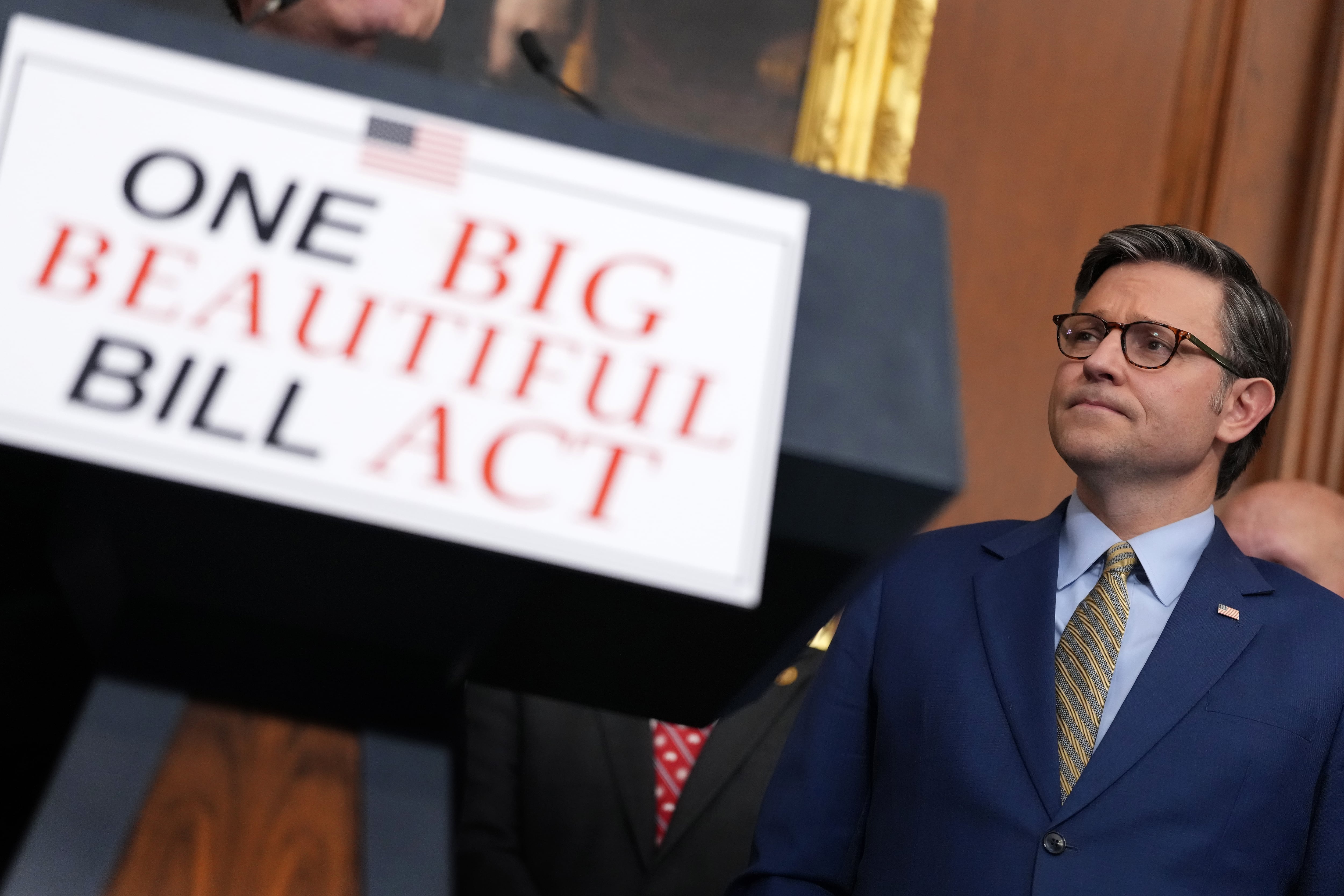 House Speaker Mike Johnson stands in front of a sign reading "One Big Beautiful Bill Act".