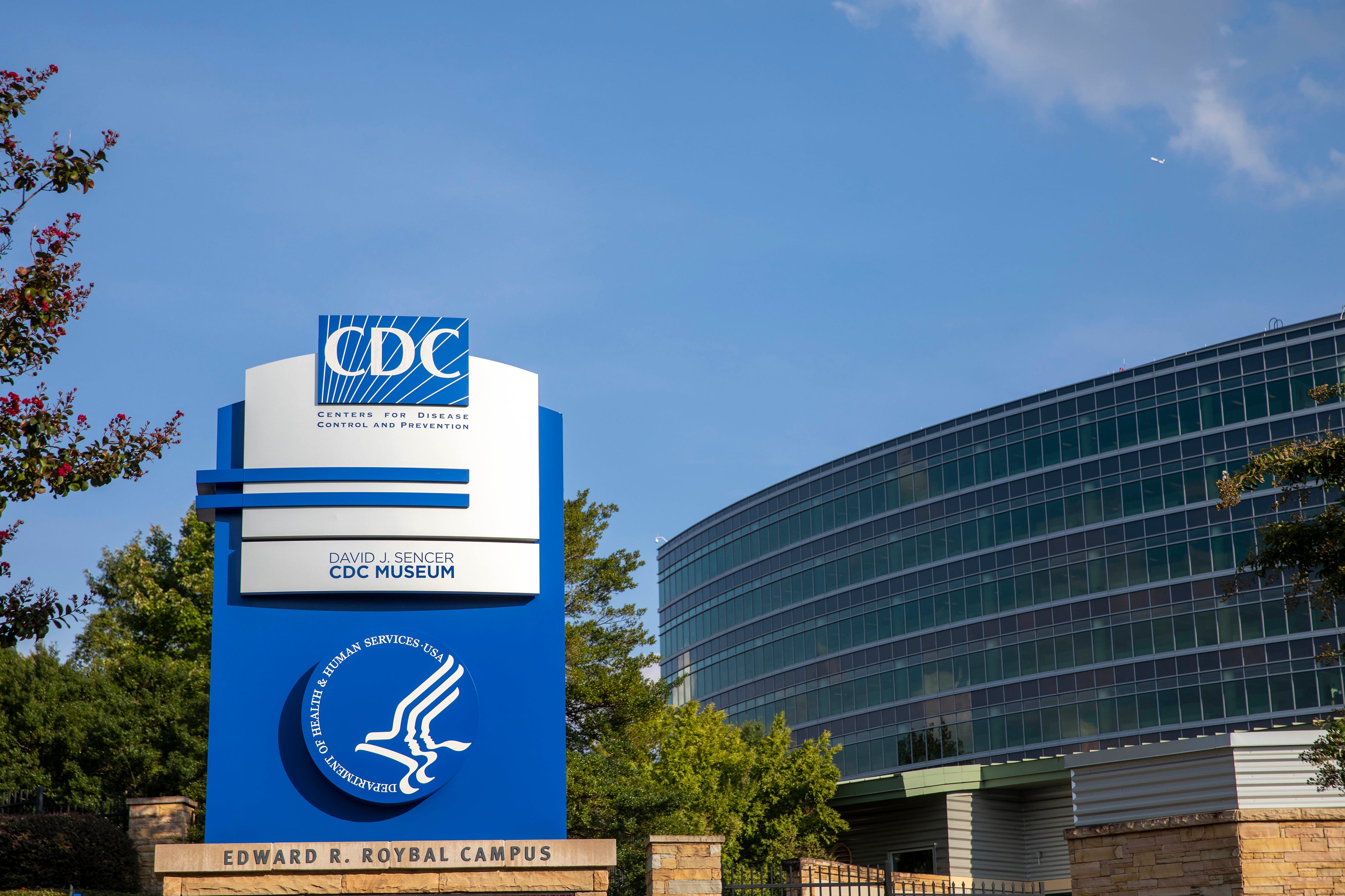 A large blue and grey sign that says CDC and other words below in front of a large glass building with a blue sky and green trees in the background.