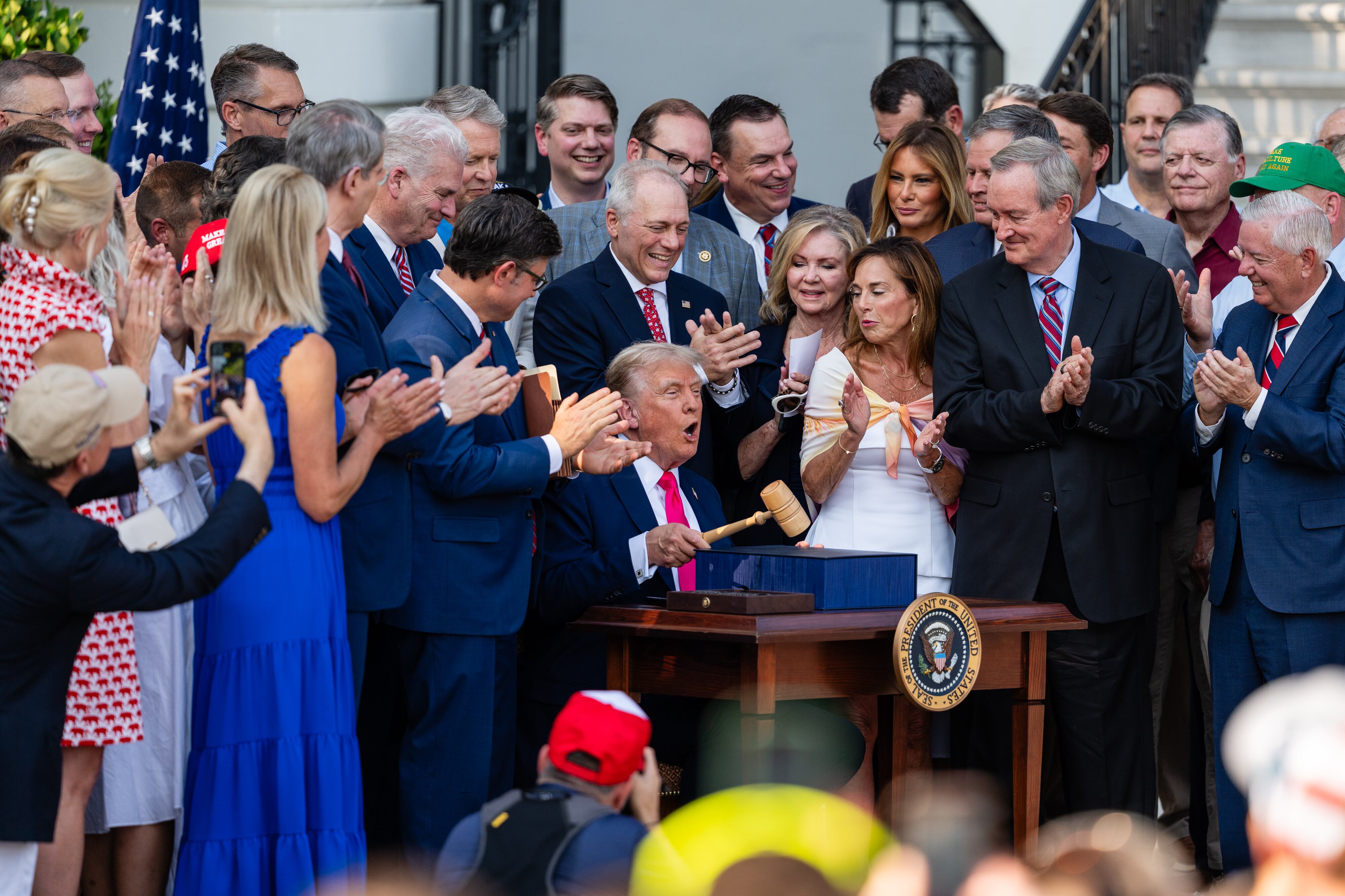 President Donald Trump sits at a desk holding a gavel, surrounded by officials, as he signs the 'One Big Beautiful Bill.'