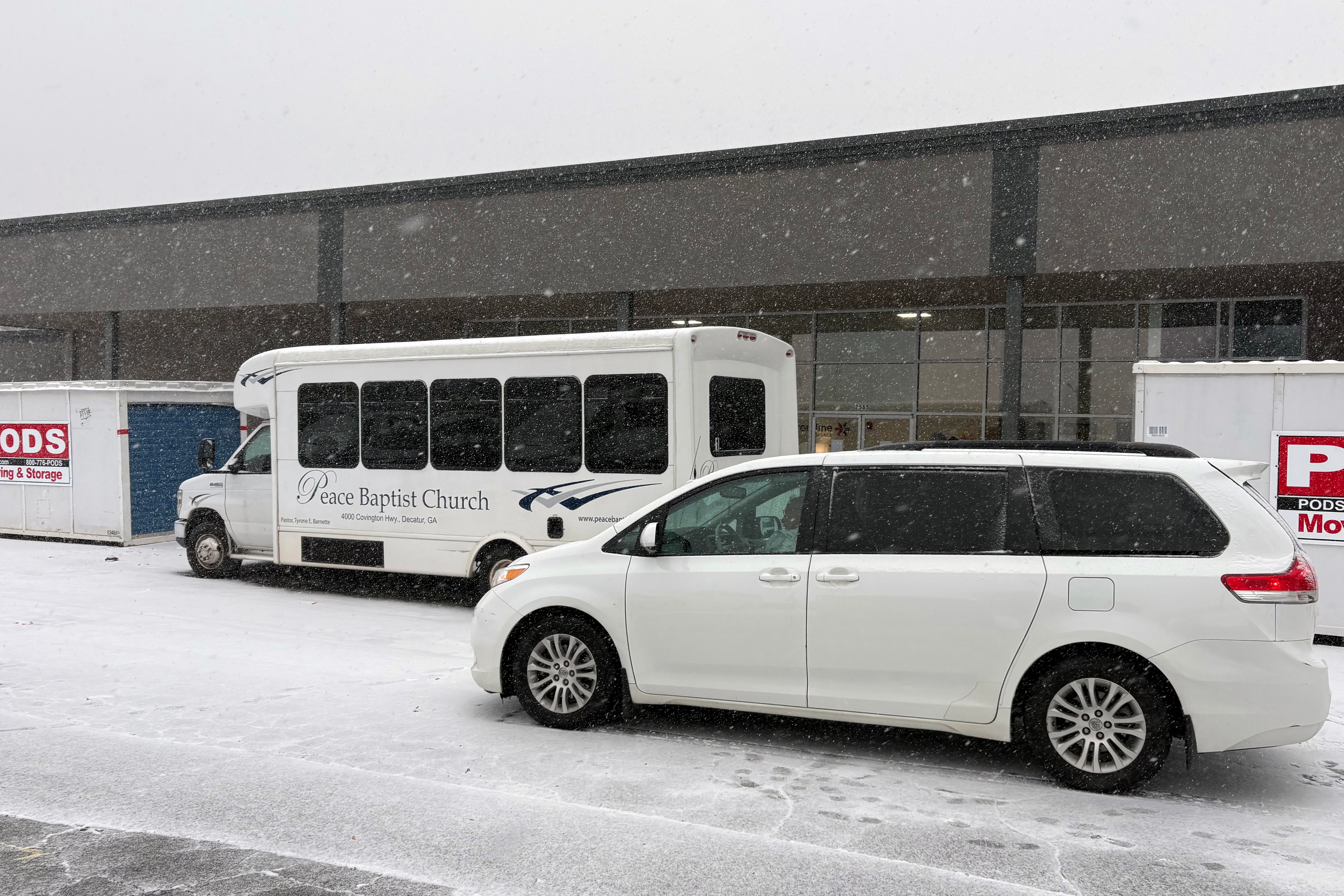 A van and a bus idle in front of a building outside with snow on the ground.