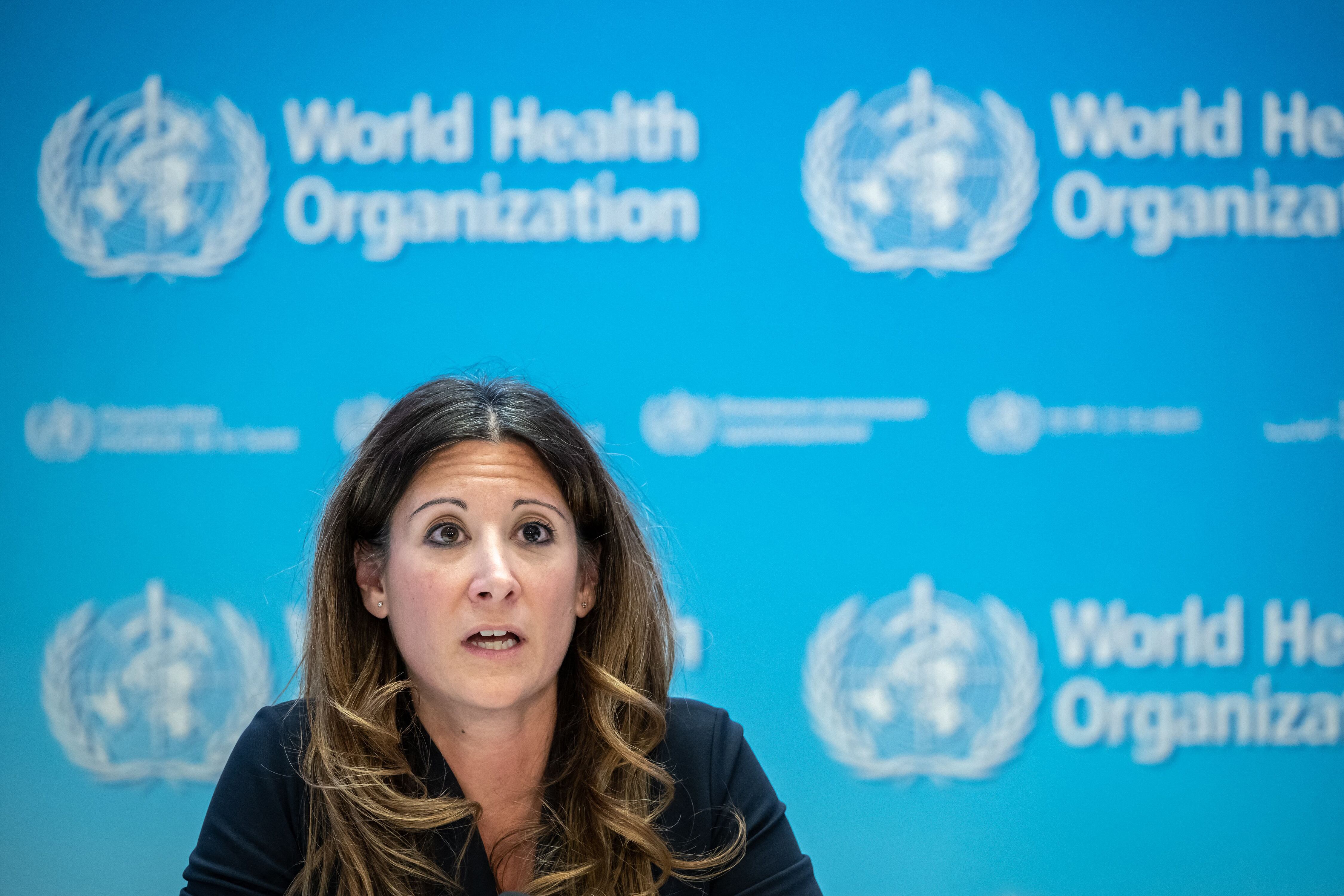 A woman with dark hair sits in front of a blue screen with white words and a logo for the World Health Organization.