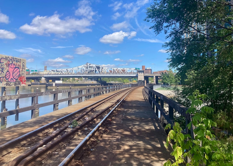 Train tracks lead your eye to a view of a city with water on the left and green trees on the right.