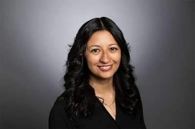 A photograph of a woman with long dark hair and wearing a black blouse posing in front of a grey background.